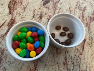 Two bowls of M&Ms, one containing a variety of brightly coloured ones, and the other containing the brown ones that have been taken out.