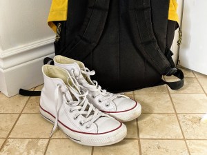 A pair of white high top running shoes next to a black and yellow backpack.